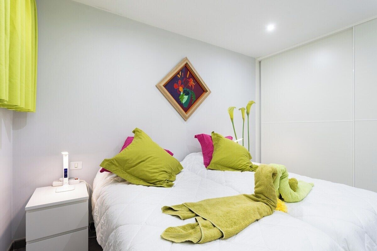 A neatly made bed with green and pink pillows, a bedside table with a lamp, and a framed artwork on the wall.