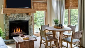 Een gezellige woonkamer met een stenen open haard, een flatscreen televisie erboven, rieten stoelen rond een houten tafel, en grote ramen met natuurlijk licht.