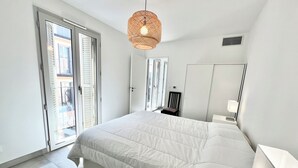 Un dormitorio con una cama grande, una lámpara colgante de ratán, una silla, una mesita de noche con lámpara y una puerta corrediza de cristal que da a un espacio exterior.