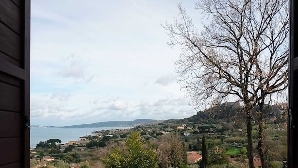 A balcony with a view of a coastal landscape, including houses, trees, and a body of water. - Breathtaking view of the lake and castle surrounded by holm oaks and oaks (Lazio)