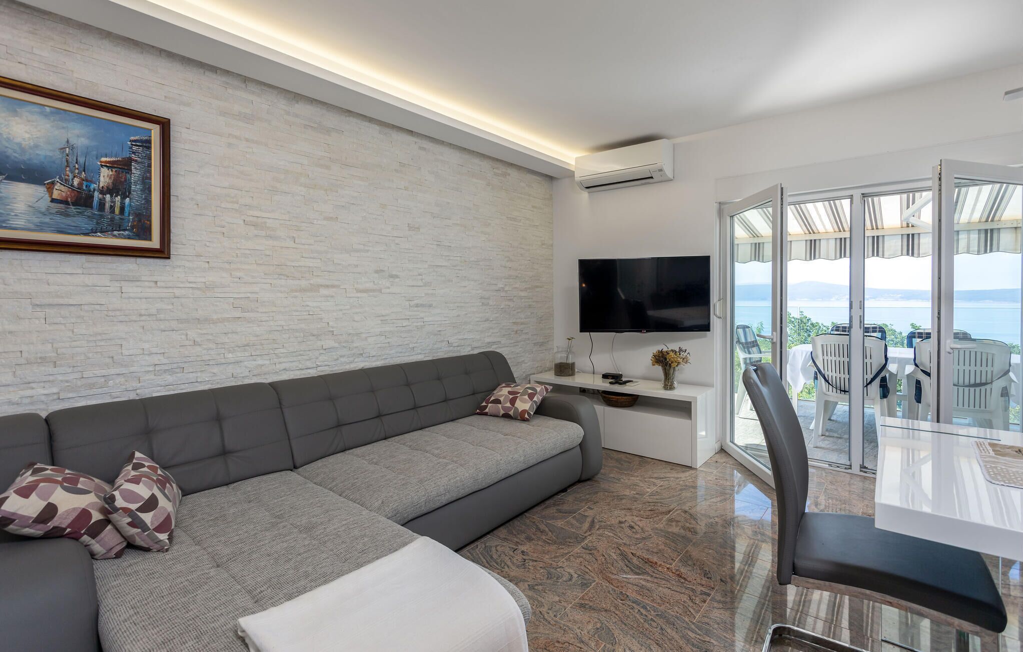 Uma sala de estar moderna com um sofá cinzento, uma televisão de tela plana e uma varanda com área de estar externa.