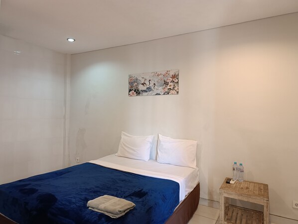 Una habitación de hotel con una cama, una colcha azul, almohadas blancas, una mesita de noche de madera con dos botellas de agua y un cuadro en la pared.