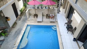 Une piscine rectangulaire entourée d’une terrasse carrelée. On y trouve deux espaces de détente en plein air, avec des chaises et des tables, ainsi que quelques plantes en pot.