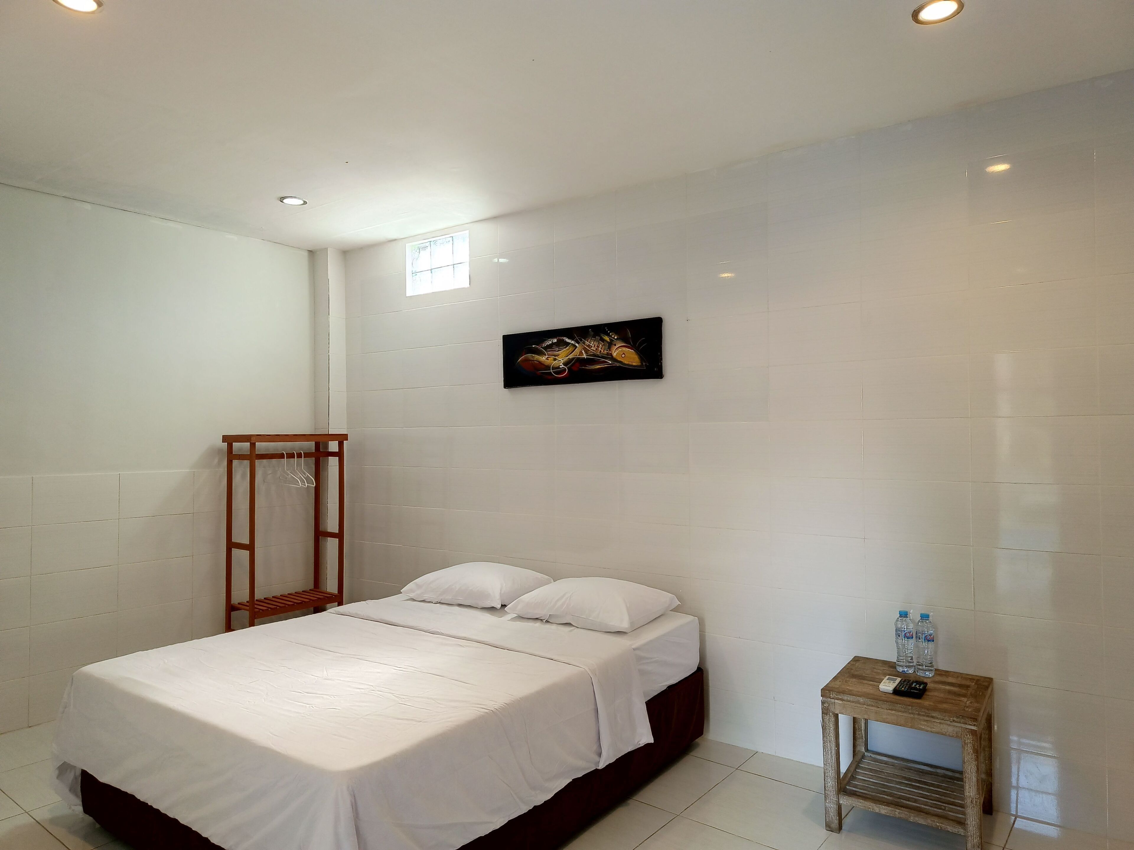 A bedroom with a bed, a wooden nightstand with two bottles, a hanging rack, and a wall-mounted artwork.