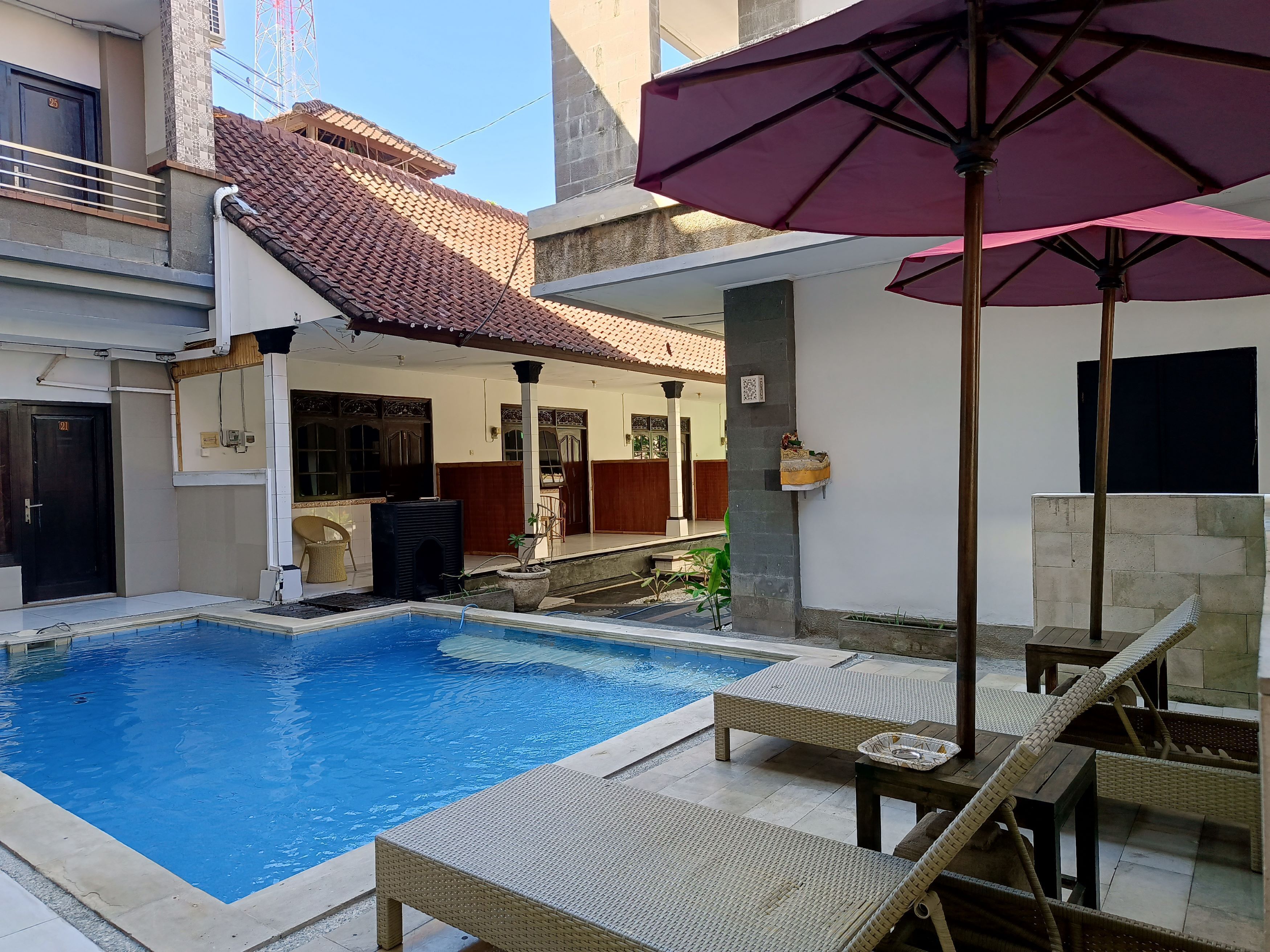 Piscina com espreguiçadeiras e um guarda-sol, cercada por um edifício com telhado telhado e uma porta com a inscrição '21'.