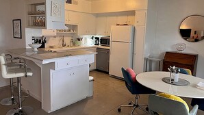 Eine kompakte Küche mit weißen Schränken, einer zentralen Kochinsel, einem Essbereich mit rundem Tisch und einem Barhocker.