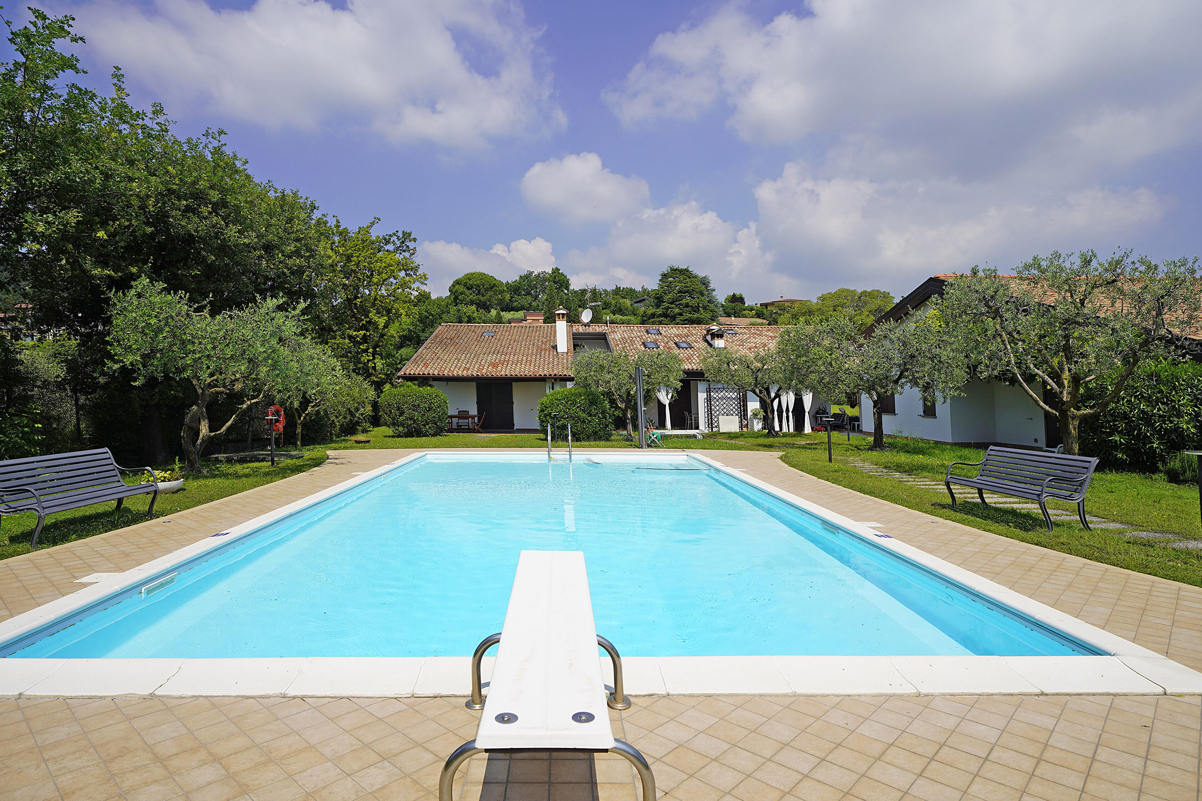 Una piscina rectangular con borde blanco, rodeada por una terraza con baldosas. Se observan bancos en la terraza, y un edificio con techo de tejas al fondo.