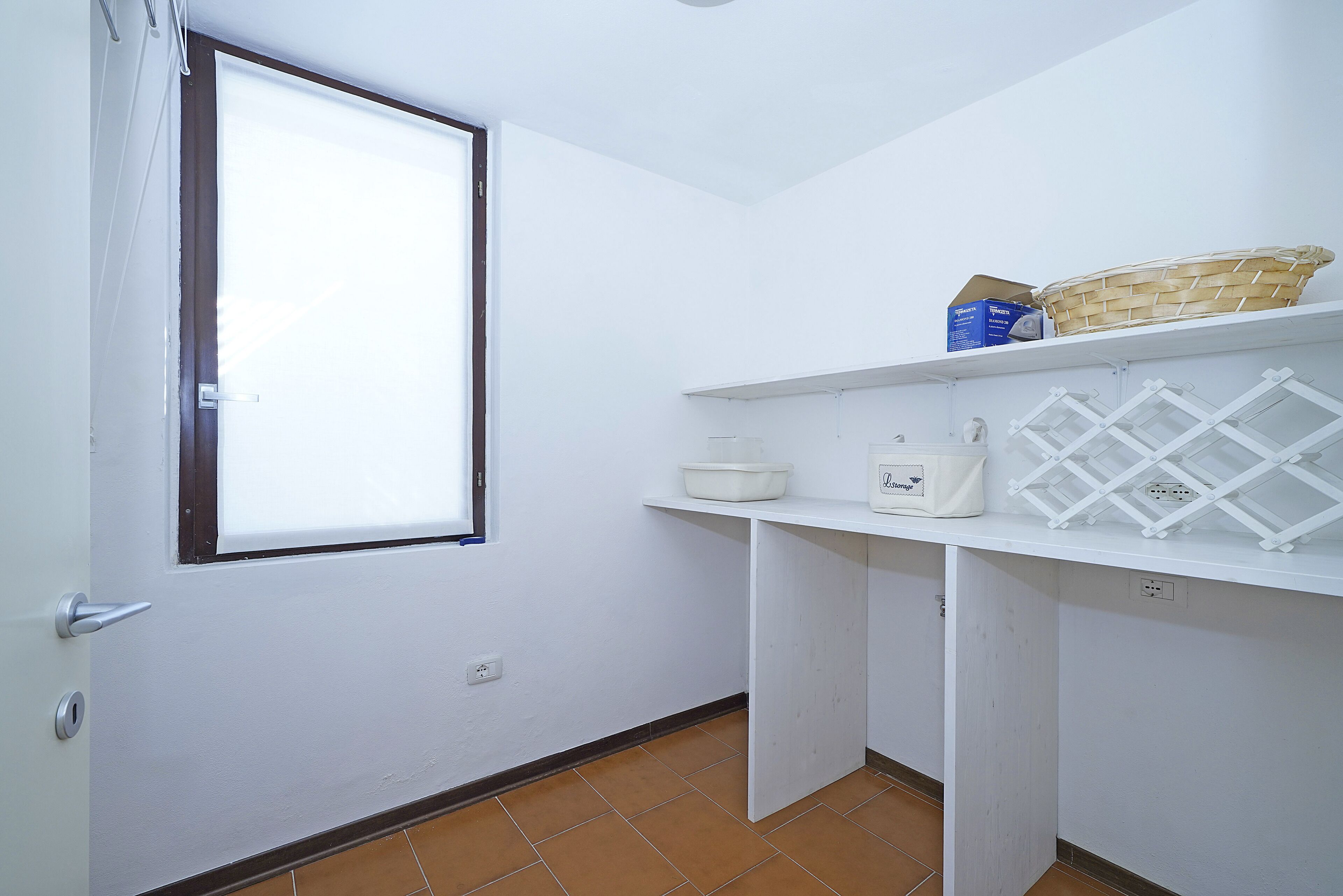 A small, bright room with a window, white walls, and a tiled floor. There are shelves with baskets and a white appliance.