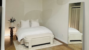 A bedroom with a white bed, a round hanging light fixture, a large mirror, and a small table with a vase.