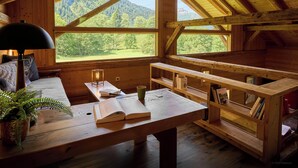 A cozy cabin with a wooden interior, a large window offering a mountain view, a table with books and a lamp, and a bench.