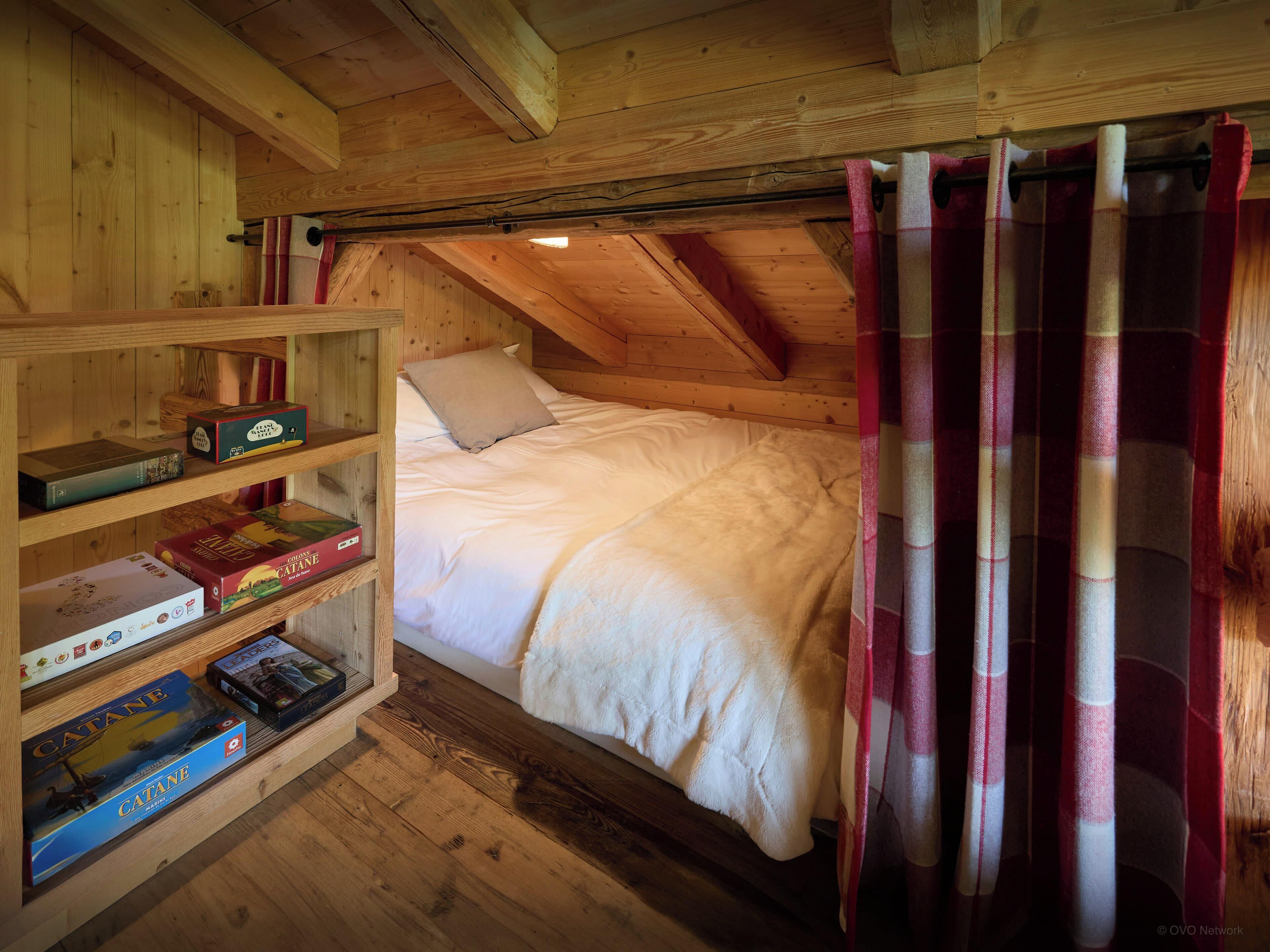 A cozy attic bedroom with a wooden ceiling, a bed with white bedding, and a shelf with board games.