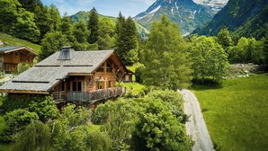 A wooden cabin with a balcony, surrounded by lush greenery and a mountain backdrop.