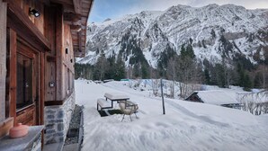 Eine Holzhütte mit Steinaußenwand, einer Bank und einem Tisch im verschneiten Hof, mit Blick auf ein Gebirge.
