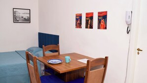 Una habitación pequeña con una cama individual, una mesa de madera con un plato y una taza, dos sillas con cojines azules, y tres cuadros en la pared.