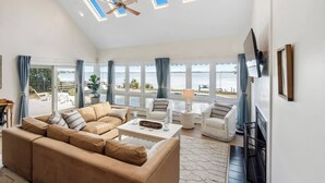 A spacious living room with a large beige sofa, two armchairs, a coffee table, and a TV mounted on the wall. Large windows offer a view of the beach and ocean.