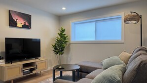 A modern living room with a flat-screen TV, a wooden entertainment unit, a round coffee table, a grey sofa with cushions, a potted plant, and a window with blinds.
