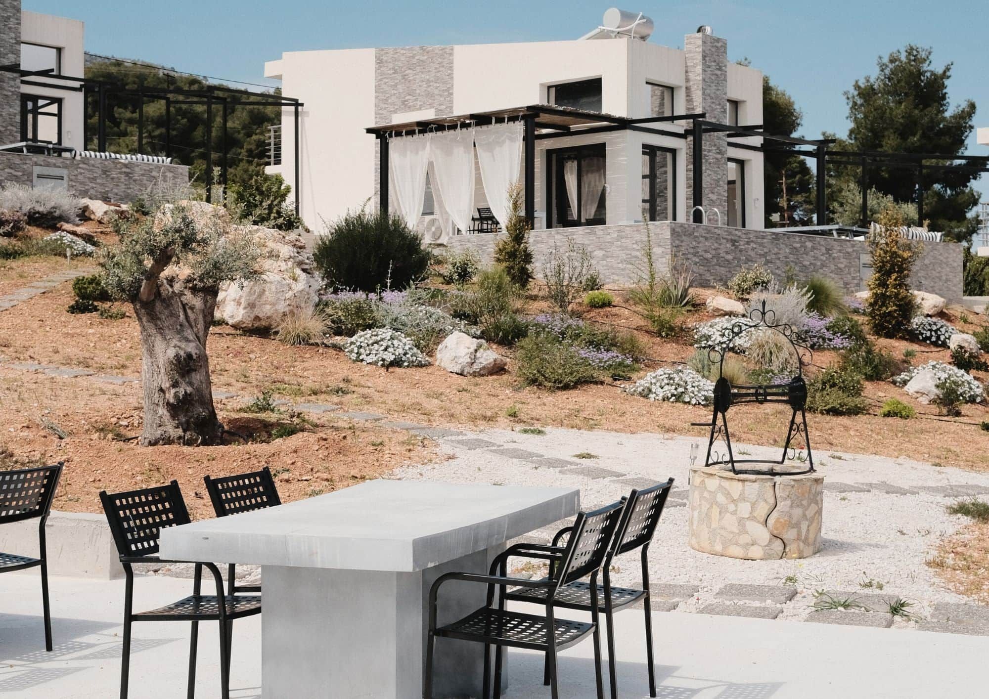 Un espace extérieur moderne, aménagé pour se détendre, avec une table en béton et des chaises en métal, entouré d’un jardin paysager agrémenté de diverses plantes et d’un grand arbre.