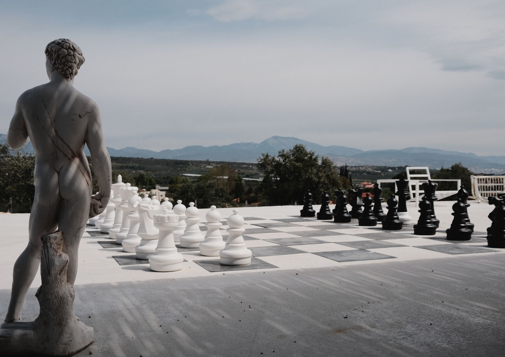 Um tabuleiro de xadrez com peças de tamanho exagerado e uma estátua de um homem em primeiro plano.