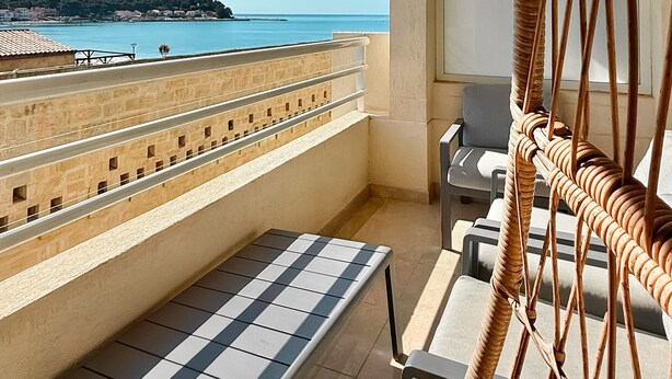 A balcony with a view of the sea, a wicker chair, and a tiled table.