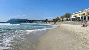Una playa con arena y olas. Hay edificios a lo largo de la costa y una montaña al fondo.