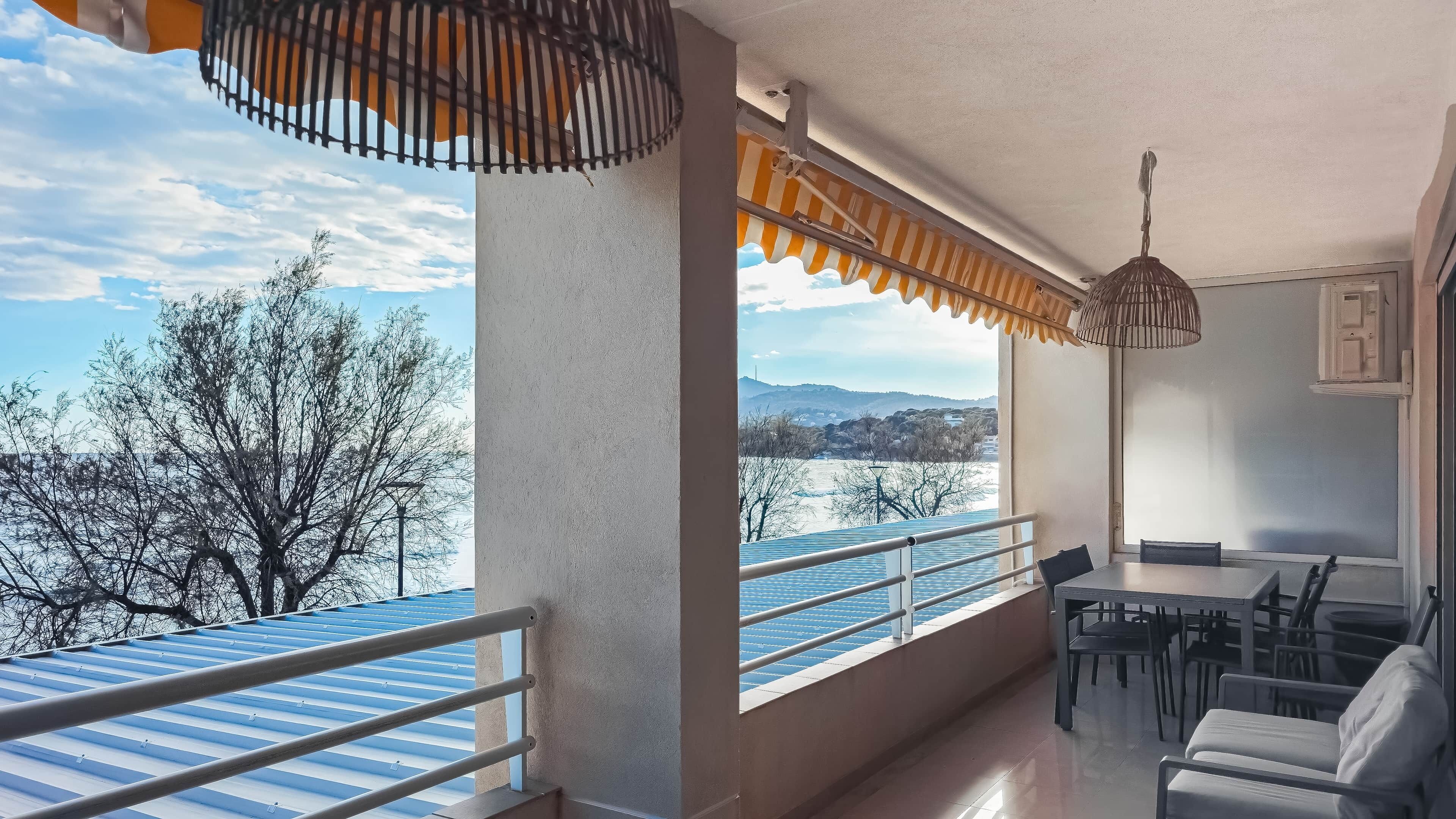 A balcony with a view of a body of water and trees, a dining area with chairs and a table, and a hanging light fixture.