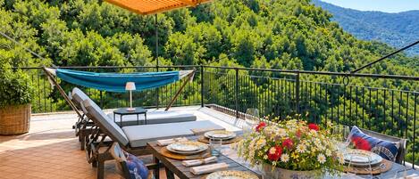 Un área de comedor al aire libre con una mesa dispuesta para cuatro personas, rodeada de abundante vegetación y montañas al fondo.