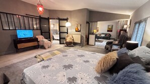 A modern hotel room with a large bed, a TV, a seating area with a chair and a sofa, and a bathroom visible through an open door.