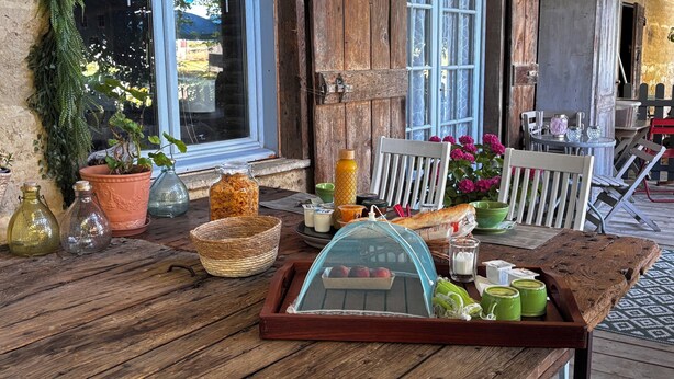 A rustic outdoor dining area with a wooden table set with food and drinks, surrounded by potted plants and a view of the outdoors.