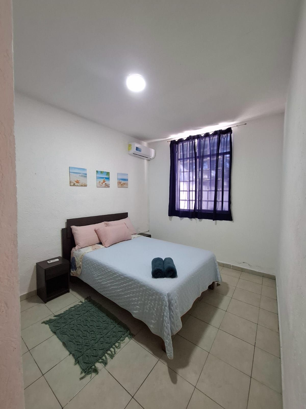 Une chambre à coucher comprenant un lit, une table de chevet, un climatiseur fixé au mur, une fenêtre avec des rideaux et trois petits tableaux décoratifs accrochés au mur.