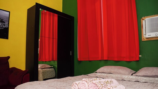 Quarto com cama, cortinas vermelhas, parede verde e um pequeno aparelho de ar condicionado.