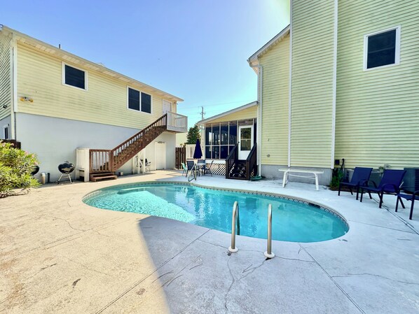 A swimming pool with a wooden deck, a covered patio area, and a staircase leading to a balcony.