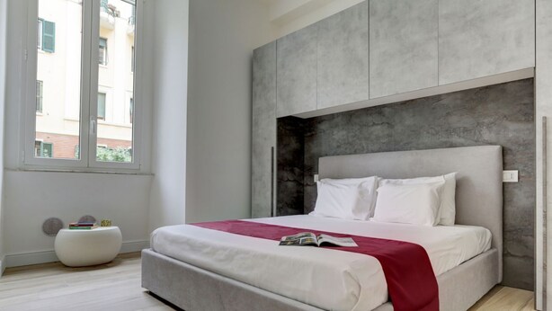 A modern bedroom with a large bed, a grey headboard, and a window with blinds. - SAN GIOVANNI COZY APT - HOME AND MORE (Roma)