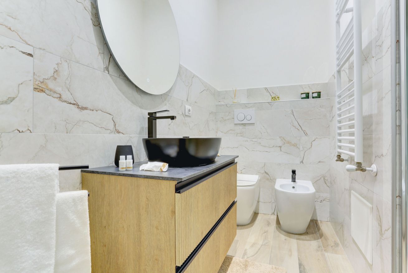 A modern bathroom with a marble wall, a wooden vanity, a black vessel sink, a bidet, and a towel rack.