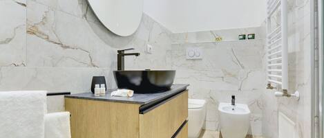 A modern bathroom with a marble wall, a wooden vanity, a black vessel sink, a bidet, and a towel rack.