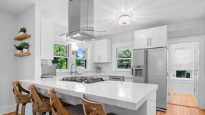 A modern kitchen with a large island, stainless steel appliances, and wooden bar stools. - Your Cape State of Mind Adventure Awaits! (South Yarmouth)