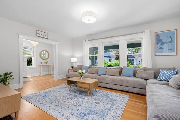 A spacious living room with a large sofa, a coffee table, and a rug. There is a window with a view of a house and greenery outside. - Your Cape State of Mind Adventure Awaits! (South Yarmouth)
