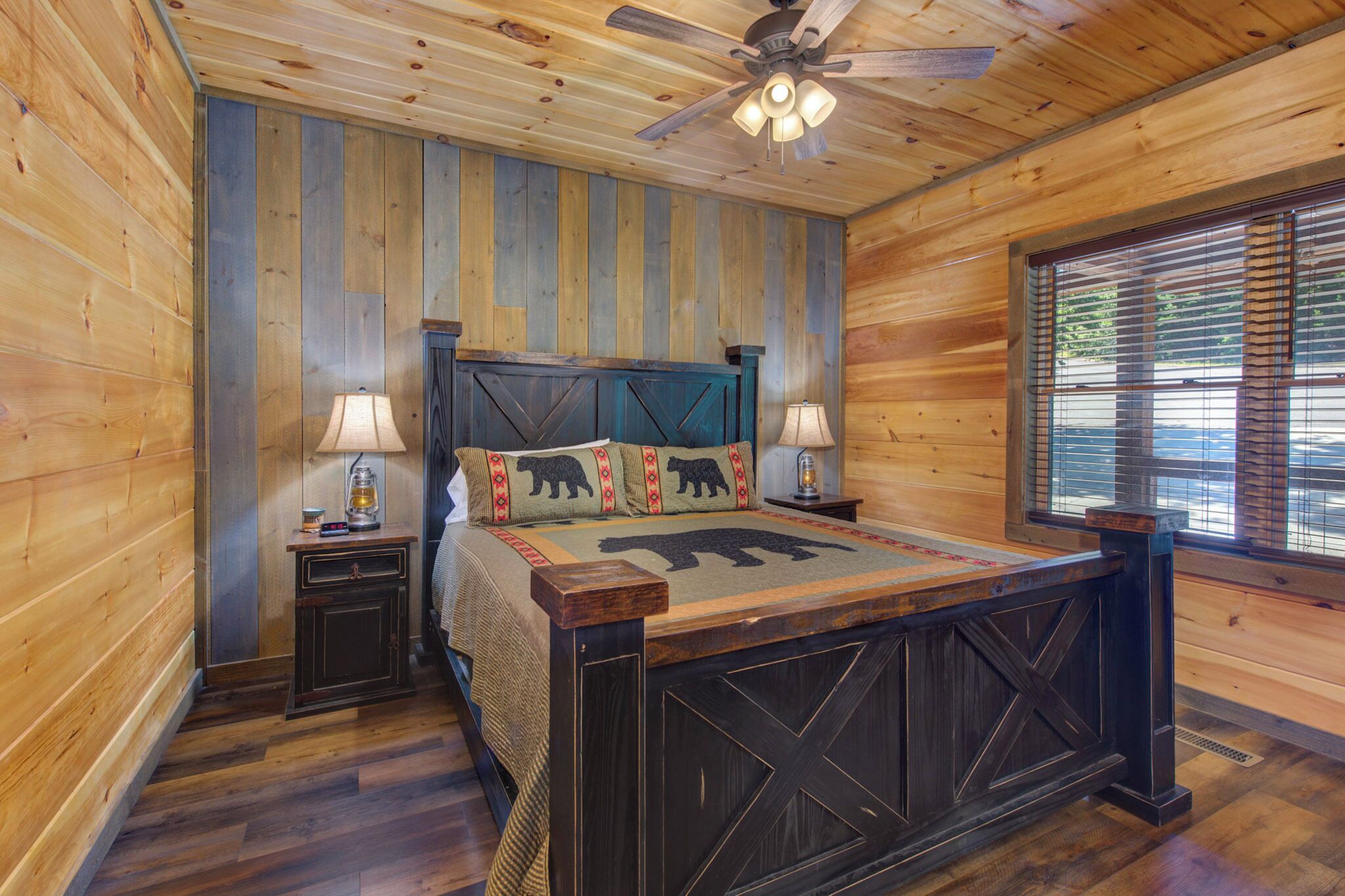 Une chambre dans une cabane en bois, avec un lit, deux tables de chevet, un ventilateur de plafond et une fenêtre avec des stores.