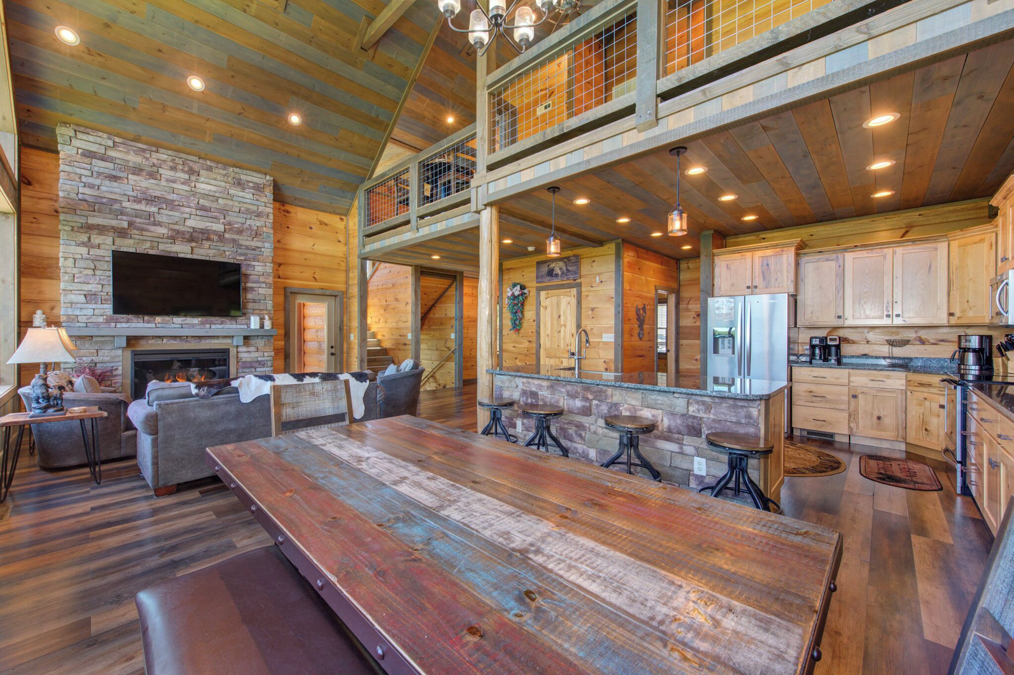 A spacious living area with a large wooden table, sofas, and a fireplace. There is a kitchen with wooden cabinets and a dining area with stools.