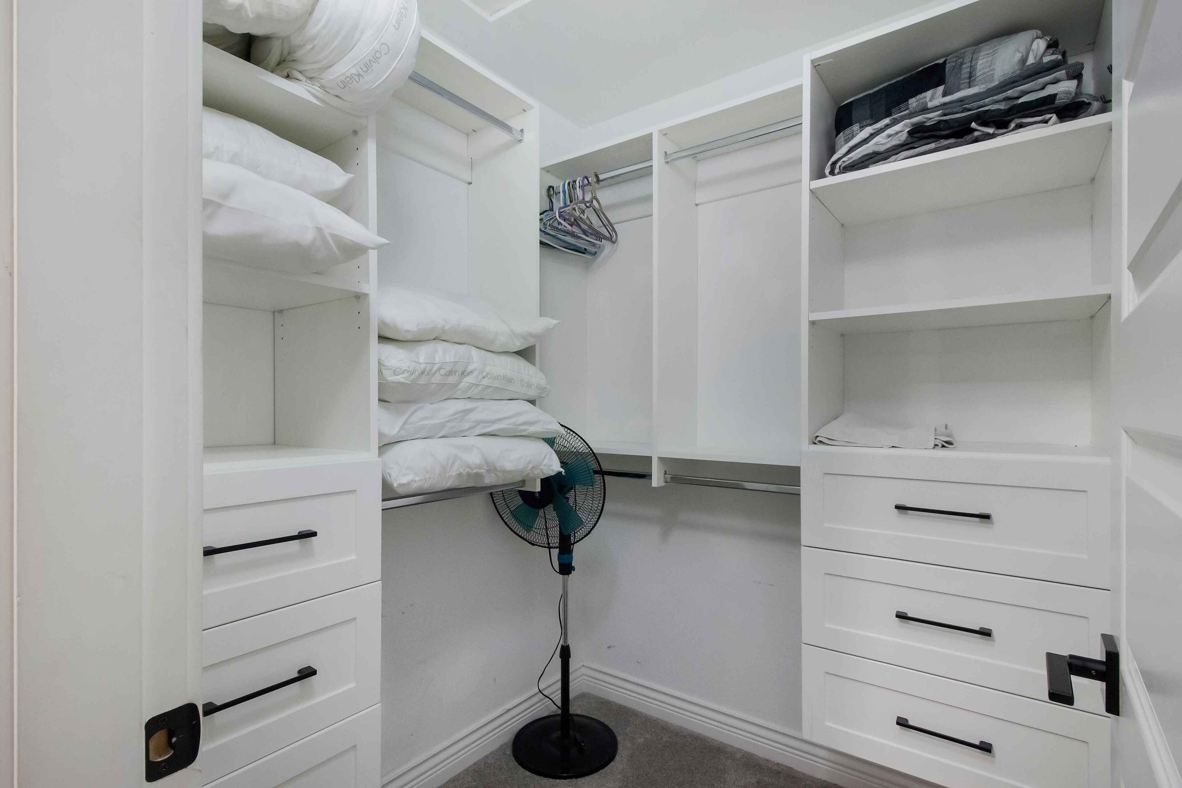 A white closet with multiple shelves, drawers, and hanging space, containing folded clothes and a standing fan.