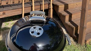 Un barbecue est installé devant une terrasse en bois, avec un bâtiment bleu en arrière-plan.