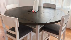 Une table de salle à manger ronde, entourée de chaises blanches, un tableau encadré au mur et une fenêtre ornée de rideaux légers.