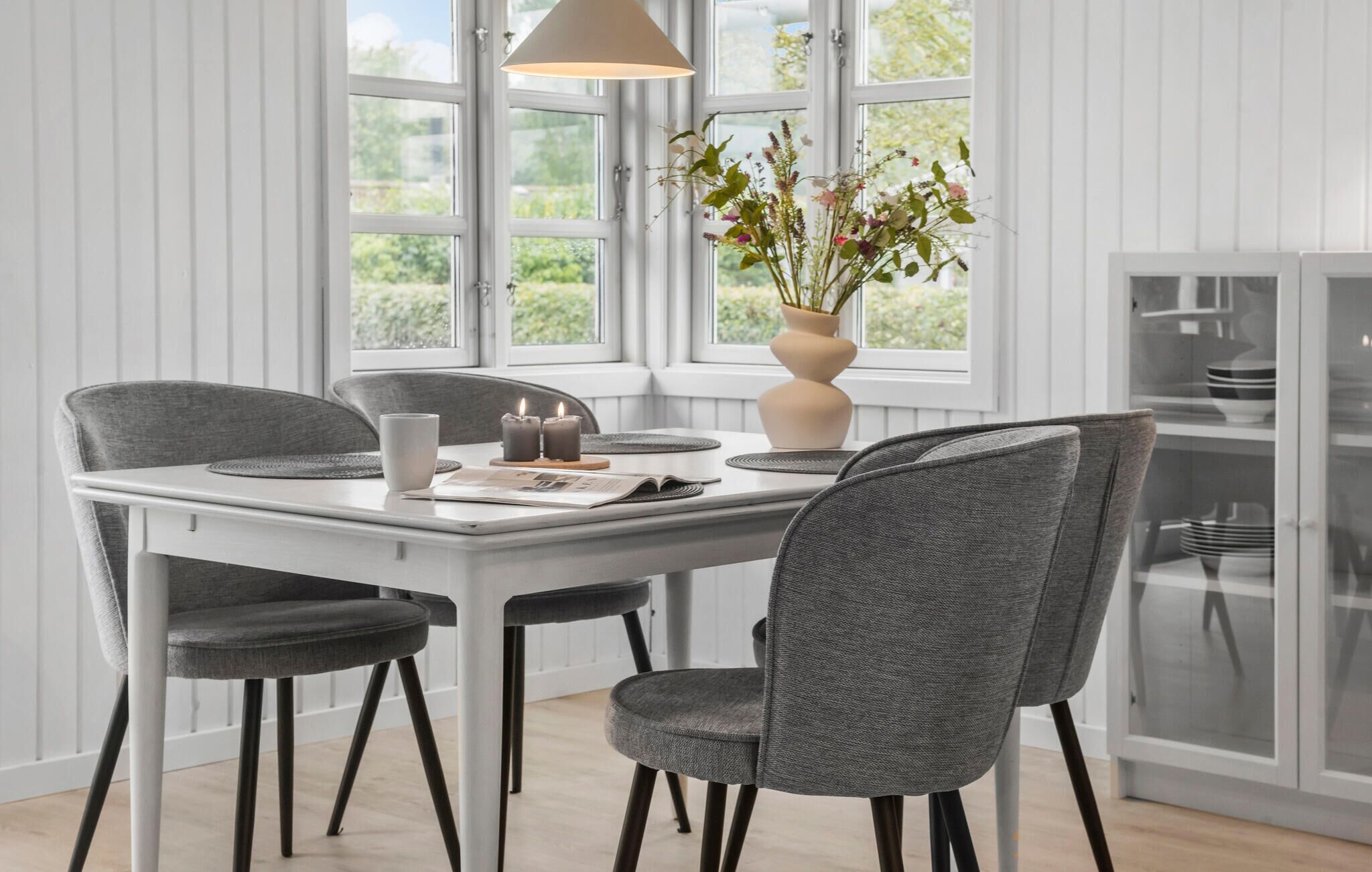 Un comedor con una mesa blanca, sillas tapizadas en gris, un jarrón con flores y una ventana con vistas al verdor.