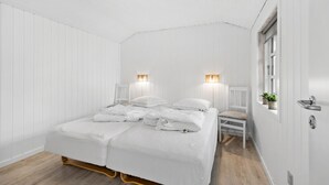 Une chambre à coucher avec un lit double, deux tables de chevet, une chaise, une fenêtre avec des plantes et une porte dotée d’une poignée.