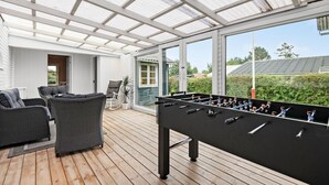 Un espace extérieur abrité comprenant une table de baby-foot, des meubles en osier et une vue sur la verdure environnante.