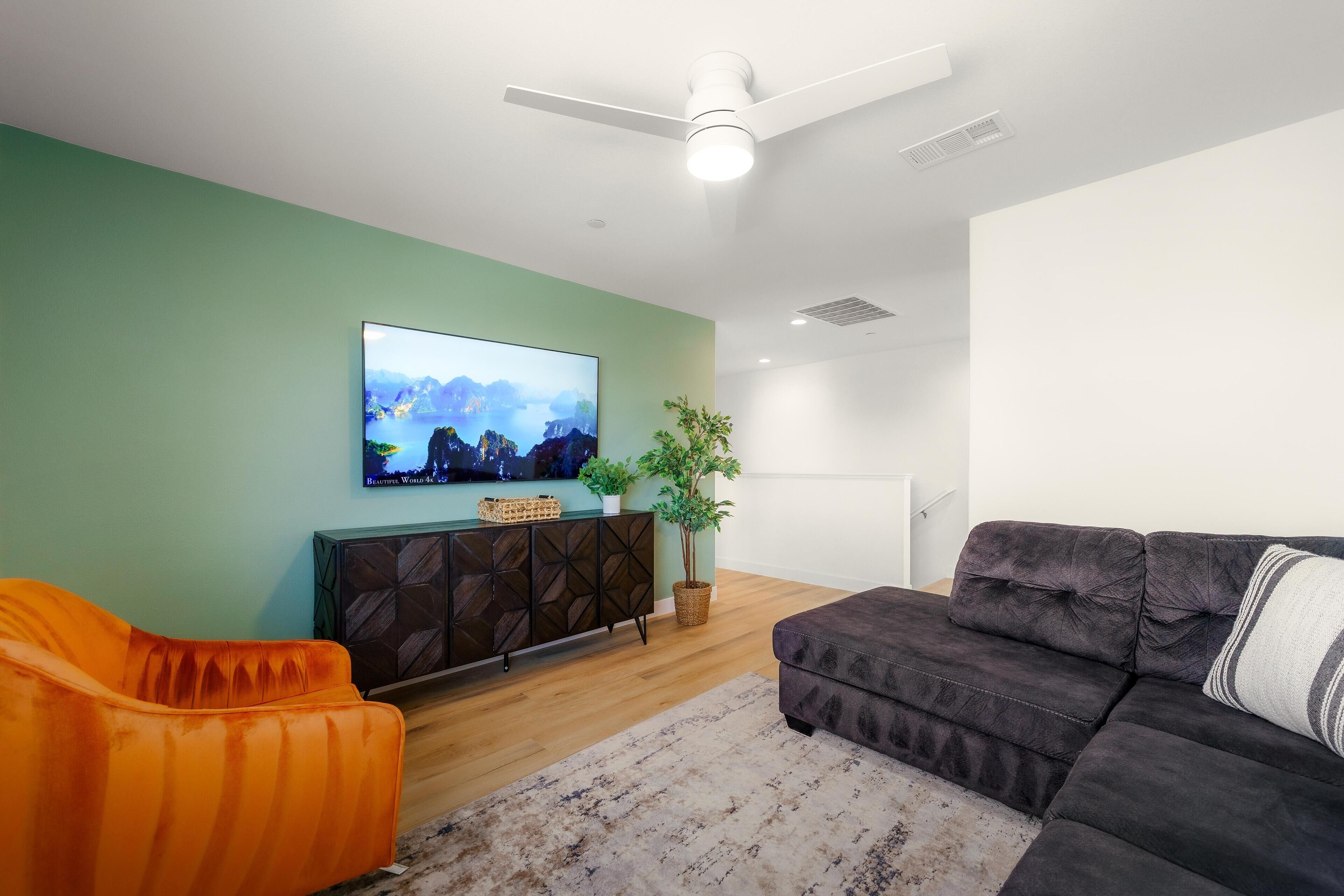 Un salon moderne doté d’un téléviseur à écran plat, d’un canapé foncé, d’un fauteuil orange, d’un plancher en bois et d’un ventilateur de plafond.