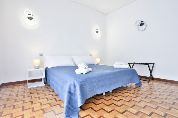 A hotel room with a bed, white nightstands, a wall-mounted light fixture, and a wooden herringbone floor.