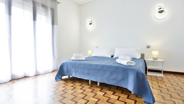 Une chambre d’hôtel avec un lit, deux tables de chevet, une fenêtre avec des rideaux et un parquet à motifs.