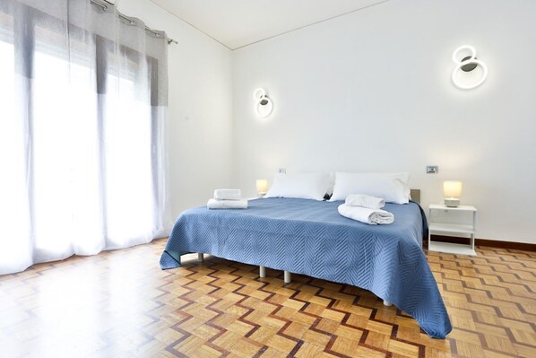A hotel room with a bed, two bedside tables, a window with curtains, and a patterned wooden floor.