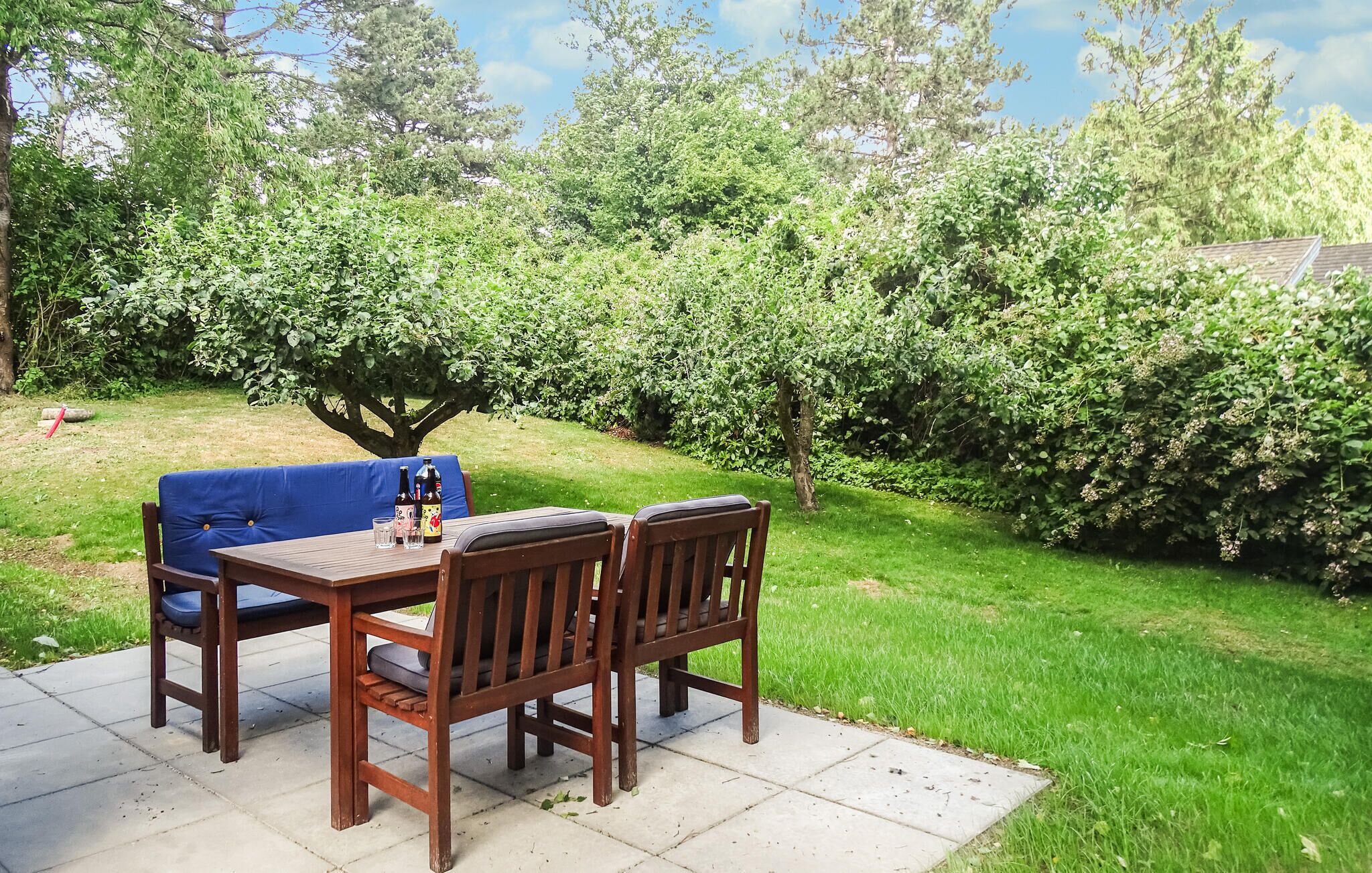 Een terras met een houten tafel en stoelen, omringd door weelderige begroeiing en bomen.
