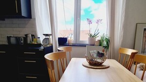Uma área de refeições com uma mesa grande, cadeiras, cozinha ao fundo e uma janela com cortinas.
